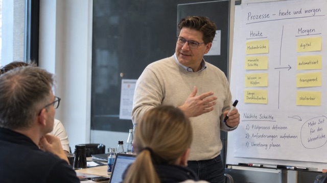 Arbeit an einer Wandtafel: Prozesse werden in zwei Spalten
                  gegenuebergestellt — wie sie heute laufen und wie sie laufen sollen.